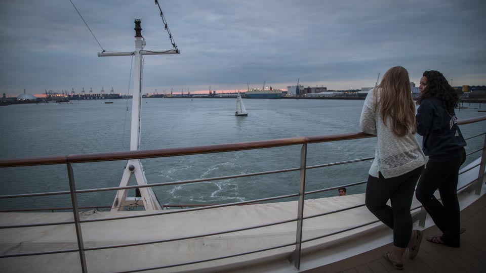 The sun sets in a cloudy sky on the shipyards in Southampton while new friends watch the city and other ship lights come on as a small wake appears behind the ship, marking the real beginning of the Summer 2014 voyage