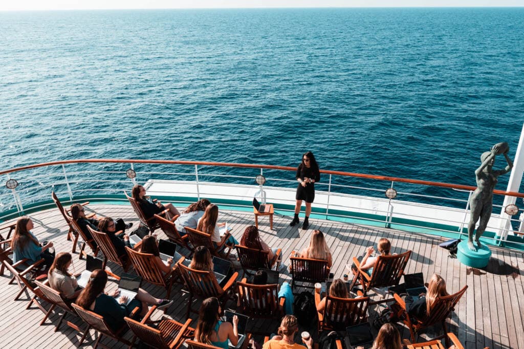 An instructor stands on the deck of a ship, speaking to about two dozen students seated in wooden chairs on the deck. Behind the instructor, the dark blue ocean stretches to the horizon.