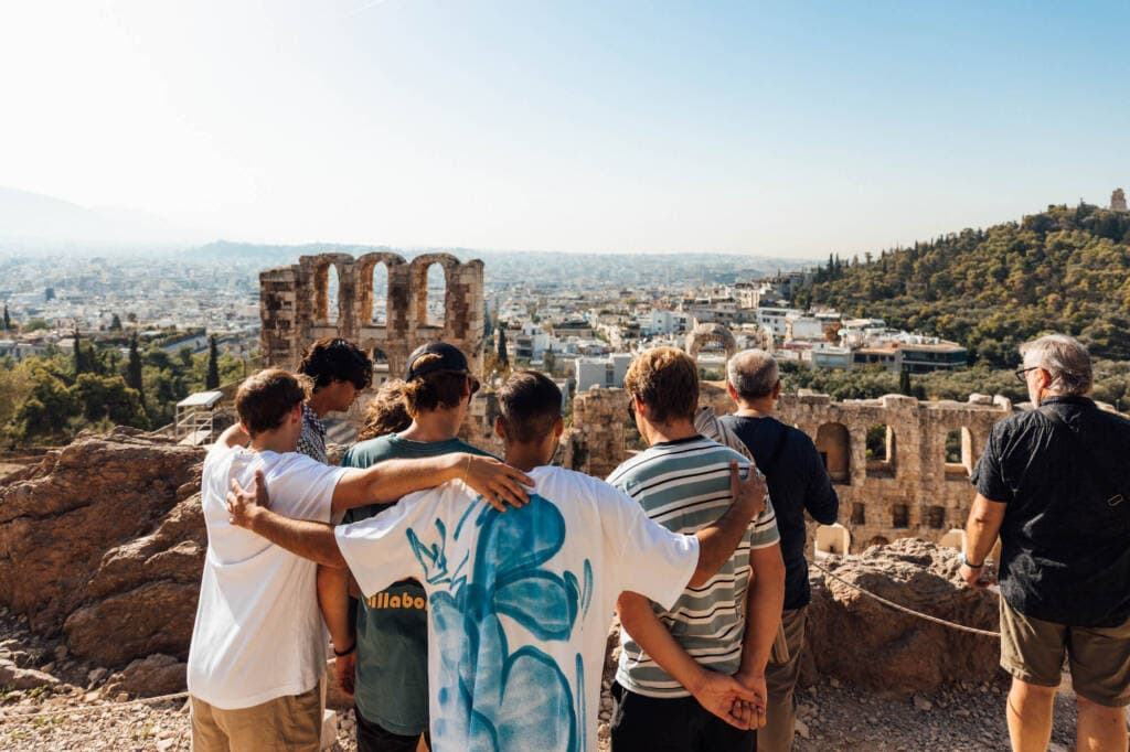 A small group of young men stand with their arms around each other and their backs to the camera, overlooking some ancient stone ruins. A city stretches to the horizon in the background.
