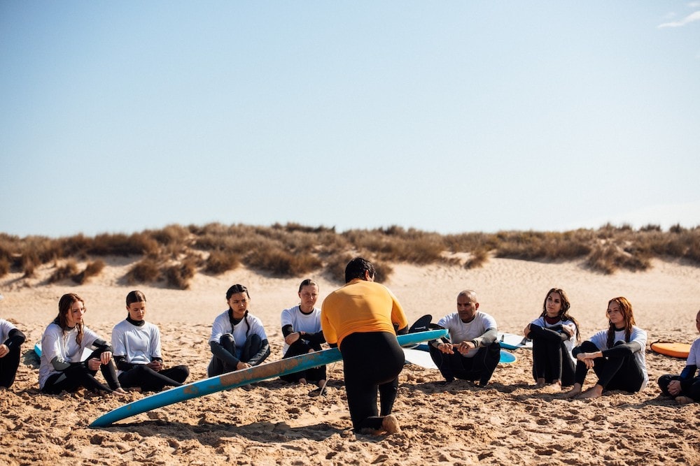 A man in a wetsuit kneels with his back to the camera and addresses a dozen people in wetsuits seated in a semicircle on a beach. The man holds a blue surfboard, and there are more surfboards visible in the sand behind the group