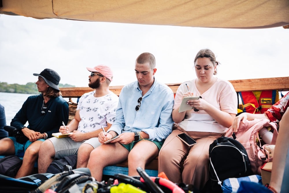 Four people sit with their backs against the side of a wooden boat, writing in small notebooks. There is diving gear visible in the foreground.