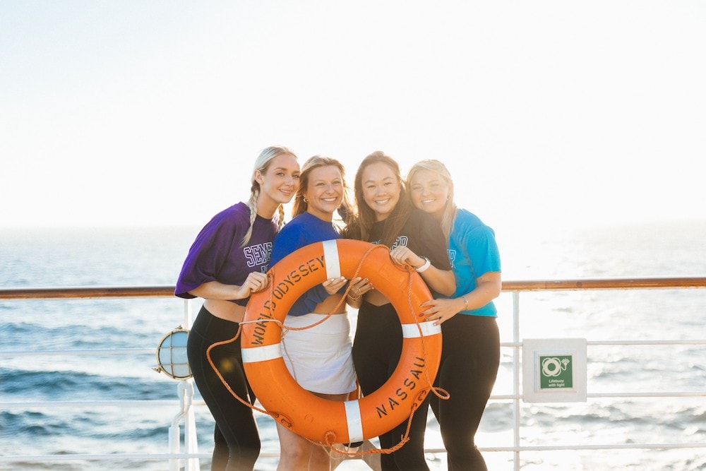 Four young women stand on the deck of a ship smiling, with the ocean in the background as they pose with an orange life preserver that reads "World Odyssey, Nassau."