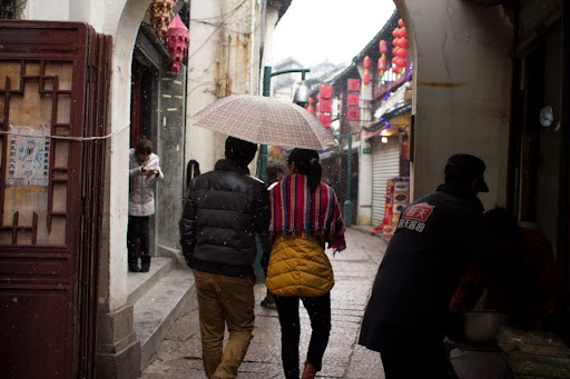 A couple in puffer jackets strolls down an alleyway, holding a plaid umbrella.