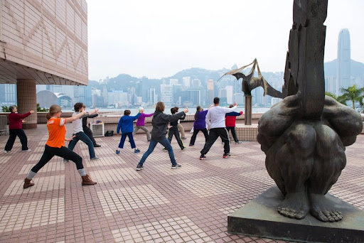 About a dozen people pose in unison, practicing tai chi between two large stone sculptures. A waterway and skyscrapers are visible in the hazy background.