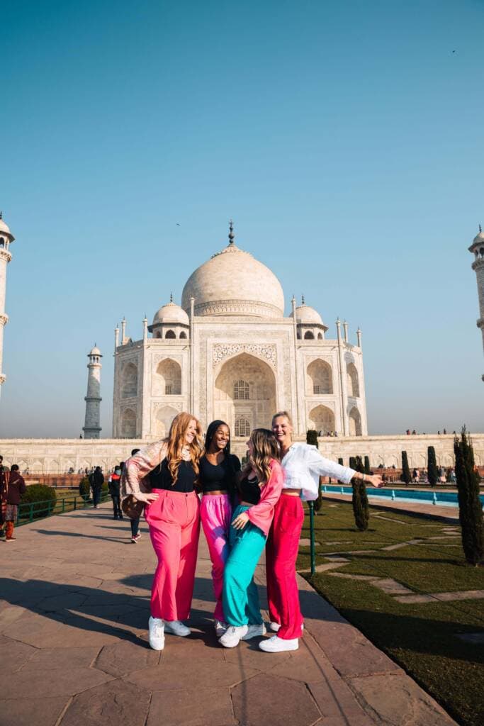 Four women in brightly colored pants and pashmina scarves laugh as they pose in front of the Taj Mahal, a giant white palace with ornately carved minarets.