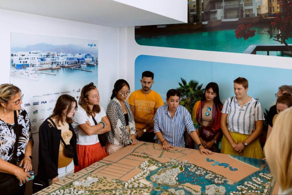 A group of people stand around a 3D tabletop model of a coastal city. A woman in the center of the group speaks and gestures to the model while the group listens.