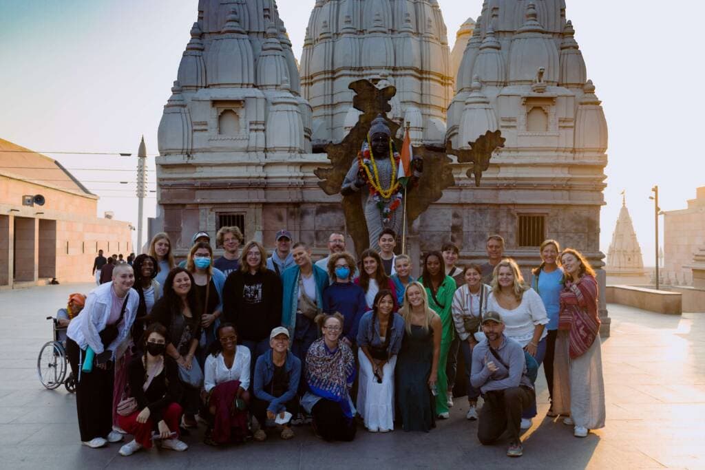 A group of about two dozen people poses in front of a life-size statue of a Hindu deity. A building with three ornately carved towers rises in the background.