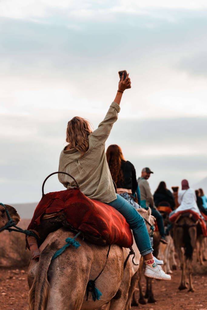 A person with shoulder length blonde hair rides a camel away from the camera while lifting their smartphone high in the air.