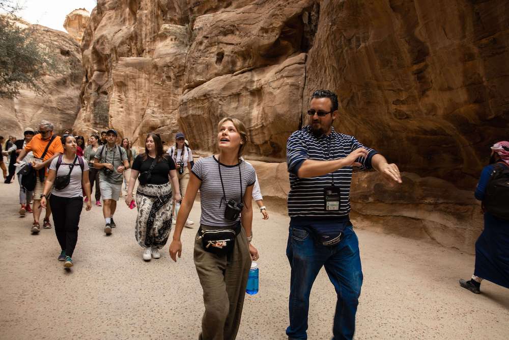 About two dozen people walk through a canyon with high rock walls. Many carry travel bags and cameras around their necks, and some gaze up at the canyon walls.