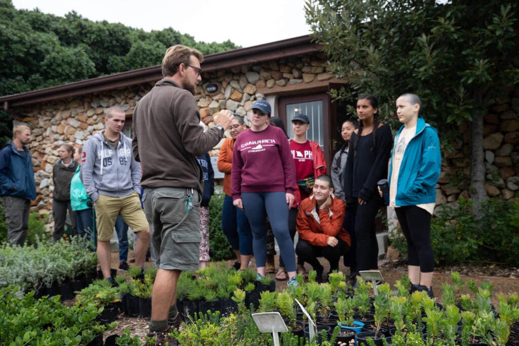 An instructor stands with his back to the camera while a group of students faces him, listening. Potted plants cover the ground at their feet.