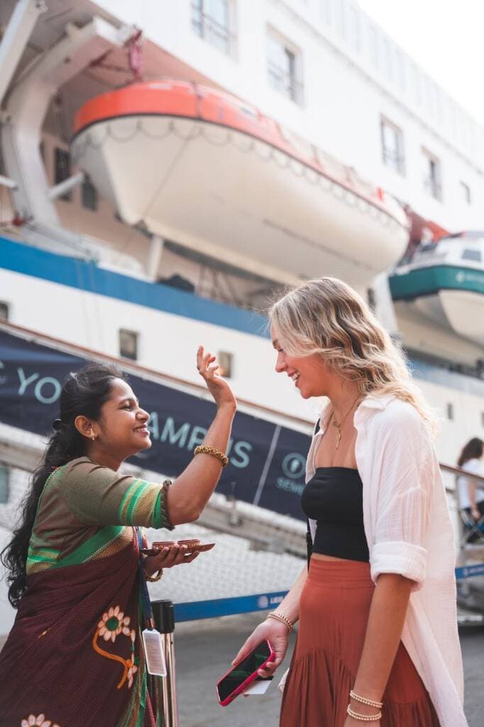 Two women face each other; one reaches out to press a finger dipped in red pigment to the other's forehead. The side of a large ship with suspended lifeboats towers in the background.