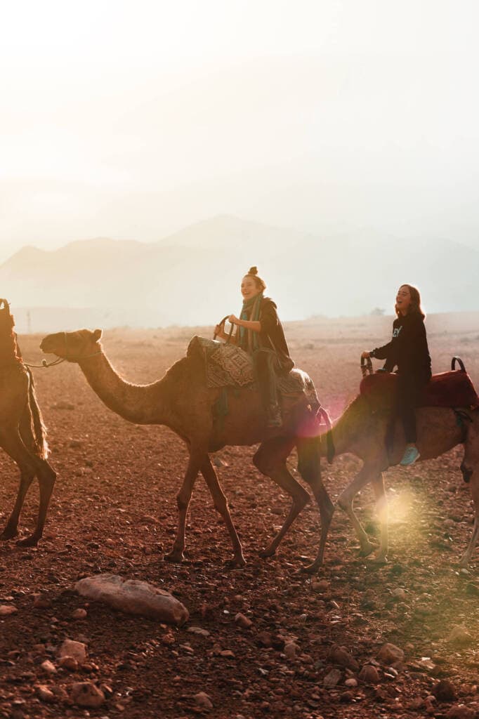Two laughing women ride camels side-by-side across a red rock desert. Hazy mountains are just visible in the background.