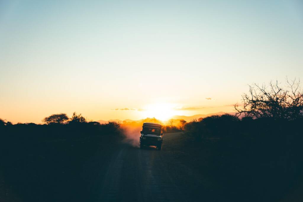 A safari jeep drives down a twilit dirt road, silhouetted against an orange sunset.