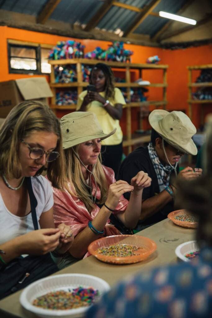 Three students sit at a table with plastic plates of beads in front of them, stringing beads on wire to make jewelry. There are shelves behind them filled with colorful animal figurines. 