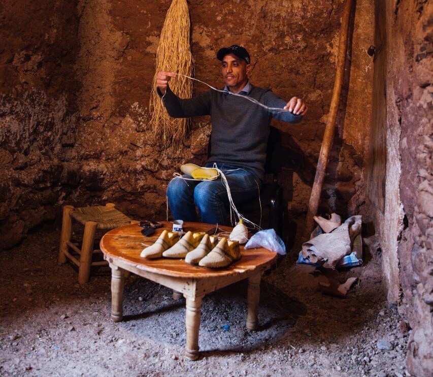 A man in a gray sweater and a baseball cap holds up a piece of twine as he balances a shoe form on his lap. A bundle of twine hangs from the stone wall behind him, while four more shoe forms sit on a low table in front of him.