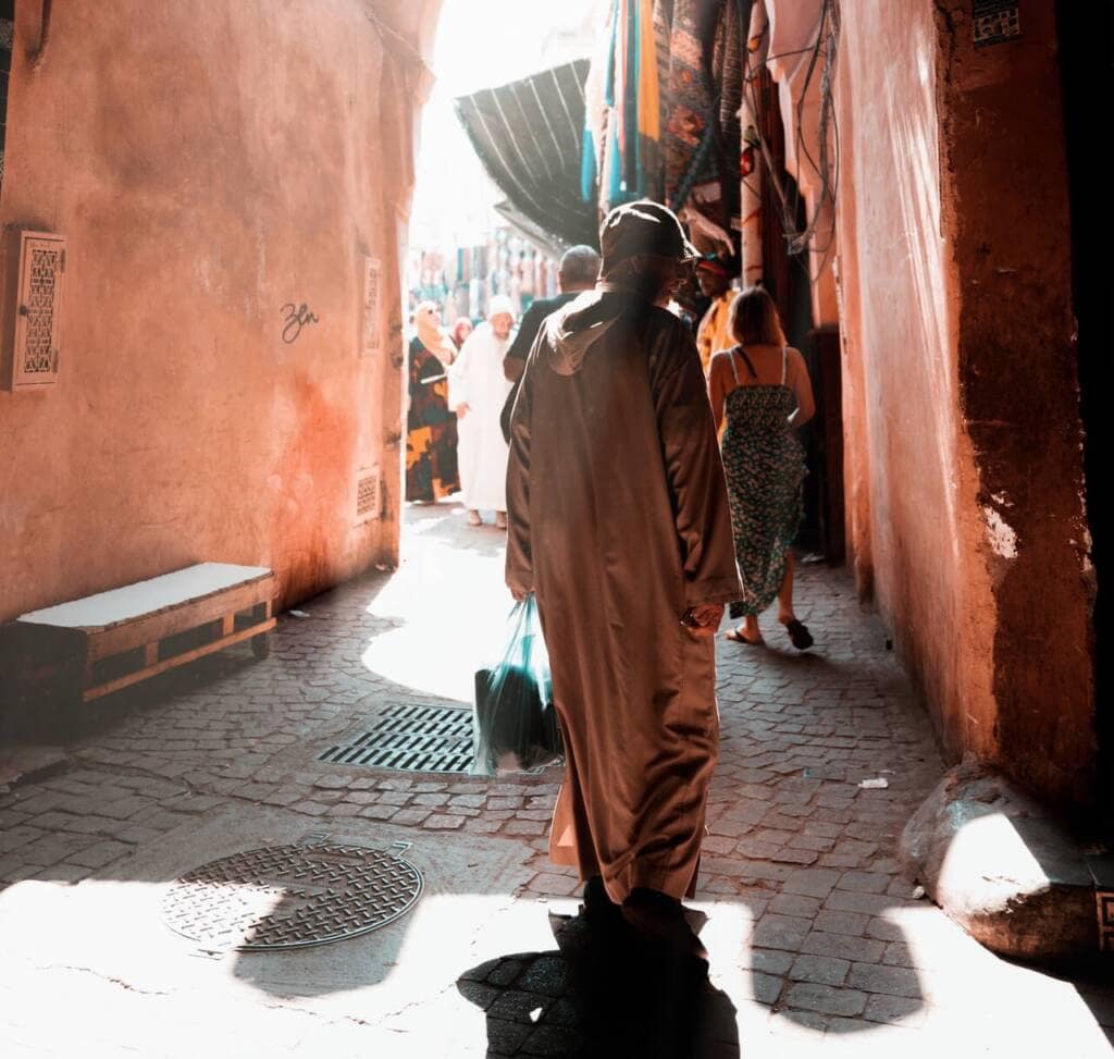 A person in a long djellaba (robe) stands in a sunlit cobblestone alleyway flanked by red plaster walls. An open-air market and shoppers are visible beyond. 