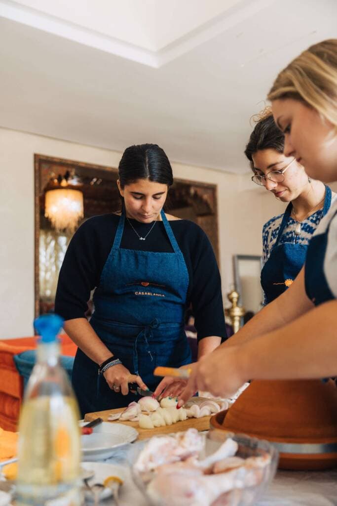 Three young women in blue aprons prepare food around a counter covered in bowls, plates, and cooking utensils. One woman chops onions on a cutting board.