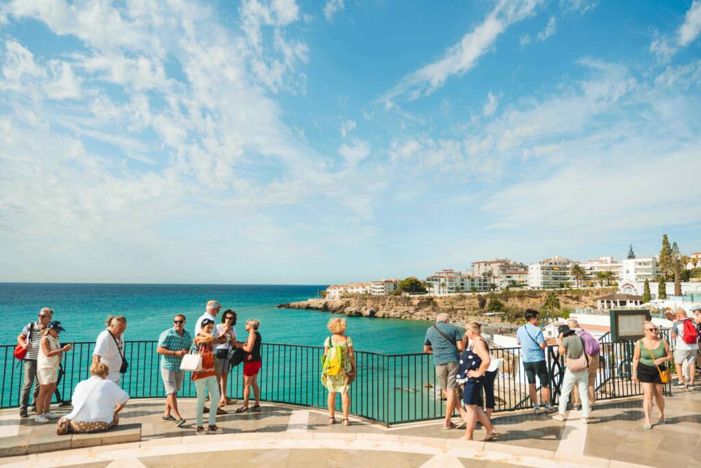 People stand and stroll on a flagstone promenade overlooking a body of turquoise water. 