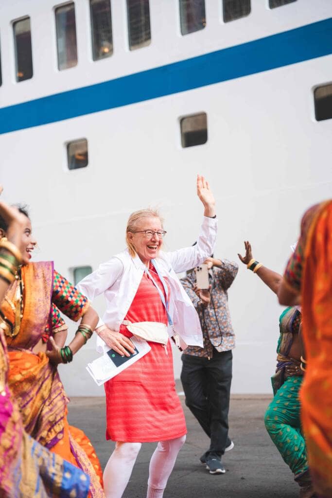 A woman with glasses and a pink dress dances among women wearing colorful saris. The side of a tall, white ship with rectangular windows provides the backdrop.