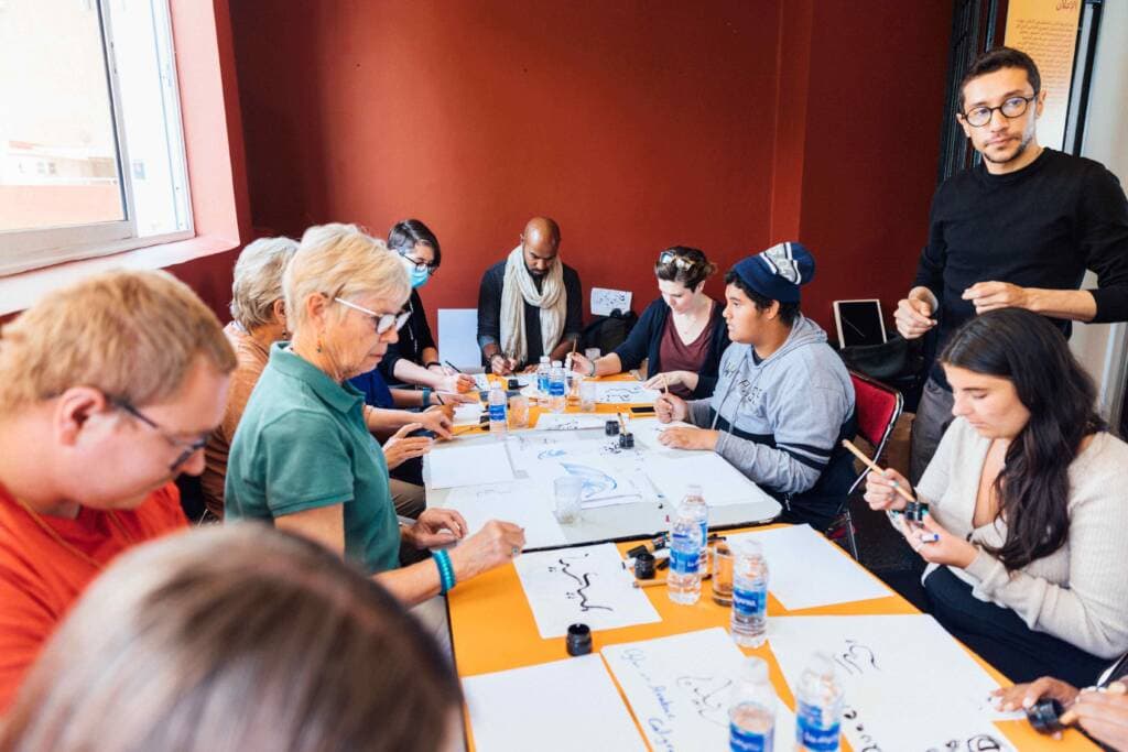 Nine people of various ages sit around a table practicing Arabic calligraphy on paper with brushes and ink. An instructor stands behind them and observes.