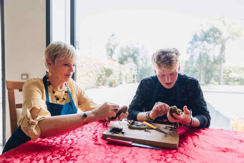 An older woman and a young man sit at a table de-seeding roasted peppers. Both wear blue aprons.