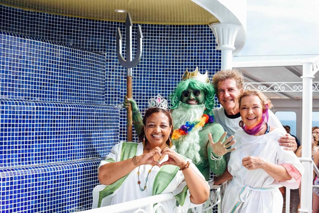 Four adults pose smiling together in front of a blue tiled wall. One is dressed as a merman, complete with green skin, a green wig, and a trident. The others are dressed in white robes; one makes a heart shape with her hands.