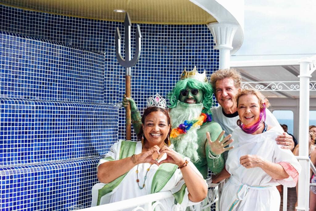 Four adults pose smiling together in front of a blue tiled wall. One is dressed as a merman, complete with green skin, a green wig, and a trident. The others are dressed in white robes; one makes a heart shape with her hands.