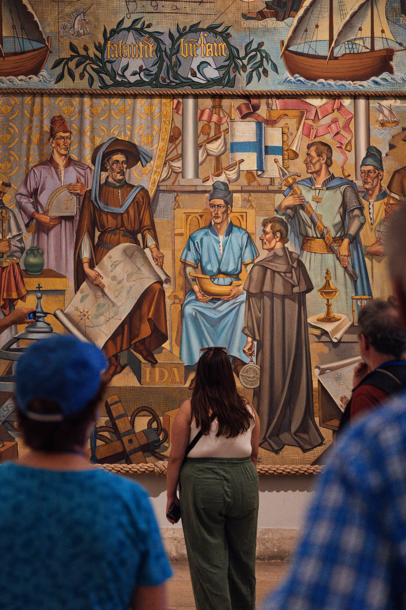 A woman in green pants and a white tank top looks at a floor-to-ceiling painting, the “Allegory of the School of Sagres,” by Portuguese painter Severo Portela Júnior. The painting depicts a half dozen men in Renaissance-era clothing holding nautical navigation objects including a sextant, map, and model ship.