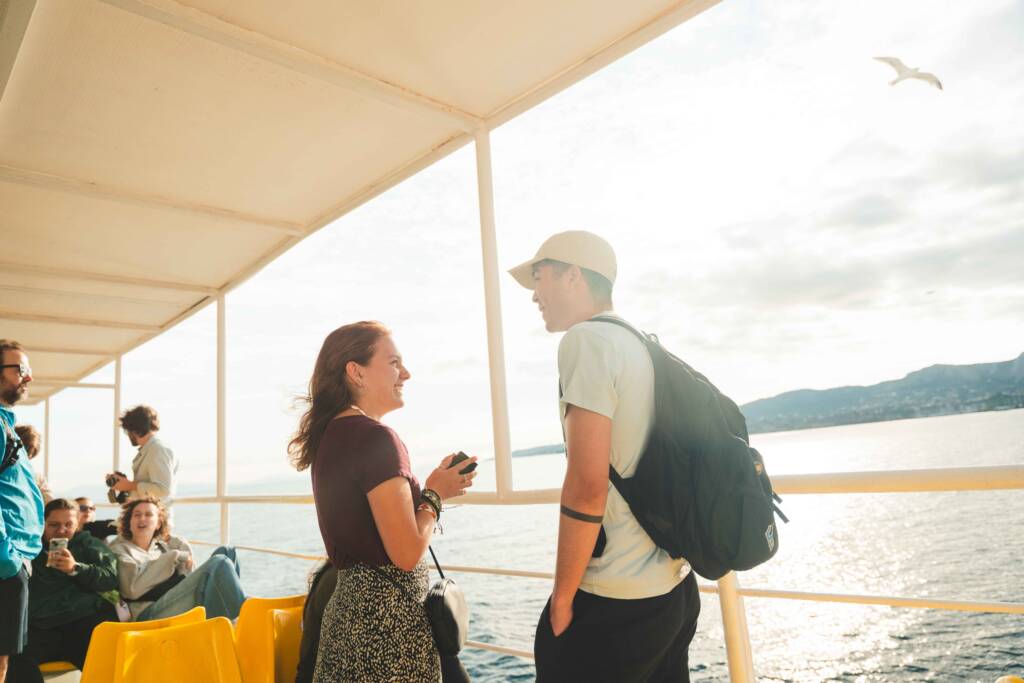 Two young people stand near the railing of a ship, talking and smiling. A sun-dappled ocean is visible beyond the railing.