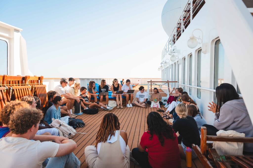 A group of about twenty students sit in a circle on the deck of a white-sided ship. Most sit on the boards, some sit on deck chairs; some have laptops open in front of them.