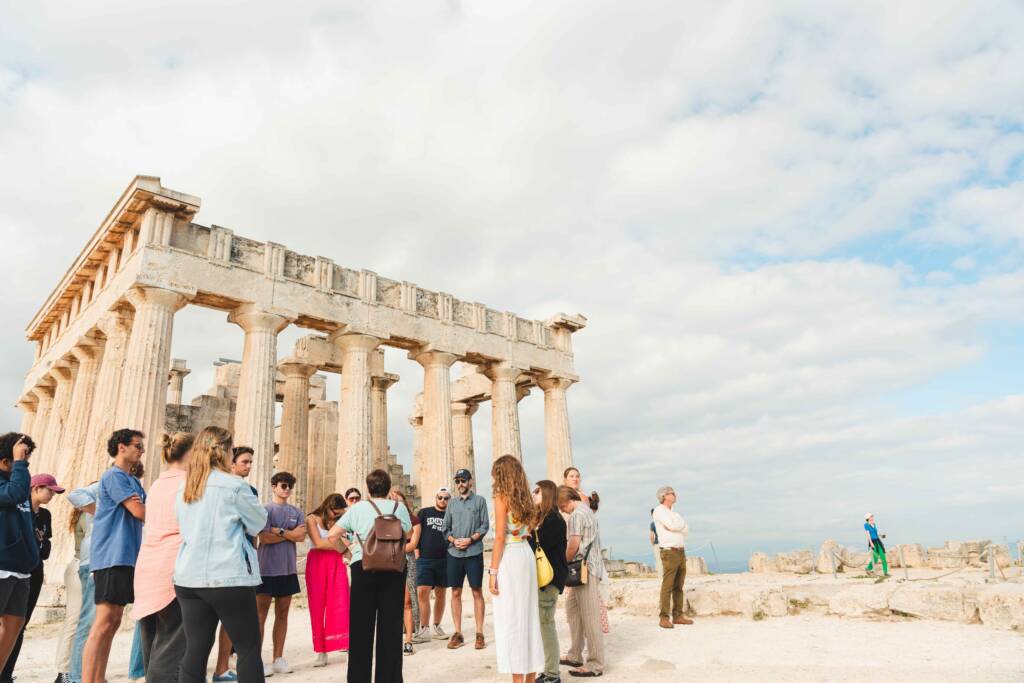 A group of about twenty people stands in front of a Roman-style ruin with pillars, listening to a tour guide.