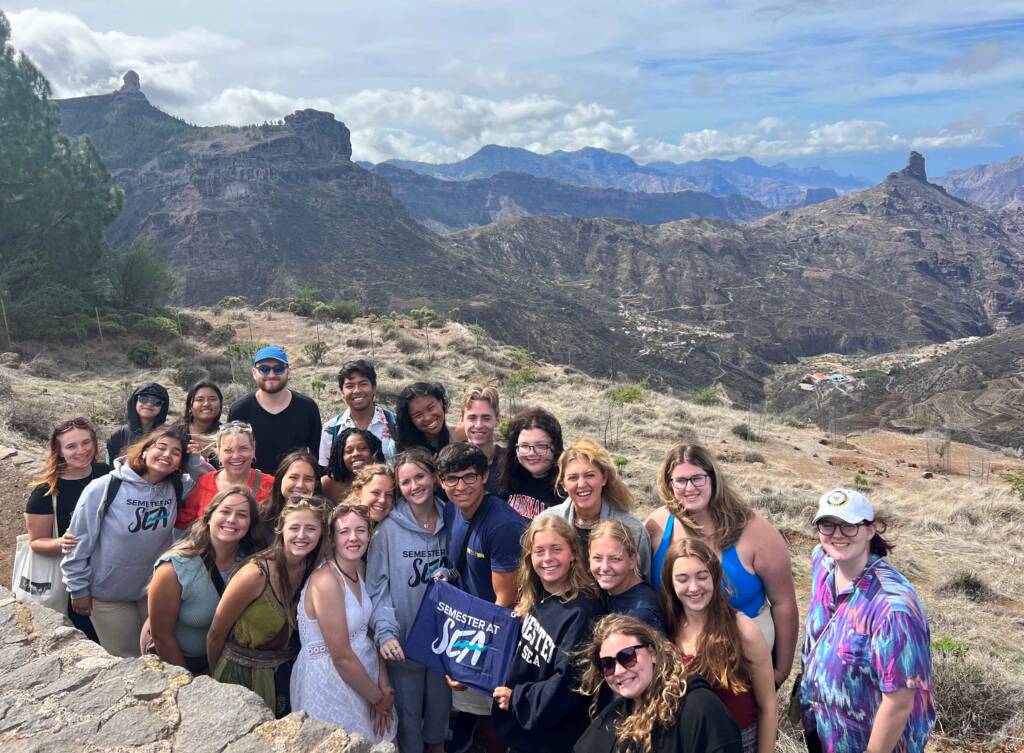 A group of about twenty-five people poses in front of a craggy mountain vista that stretches into the distance. Three people at the center of the group hold a small blue flag that reads “Semester at Sea.”