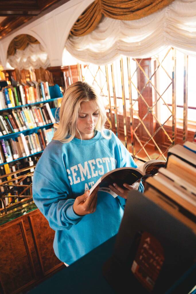 A young woman with long, blonde hair stands in front of a full bookcase, looking at an open book in her hands. She is wearing a light blue sweatshirt featuring the words “Semester at Sea” in white letters on the front.