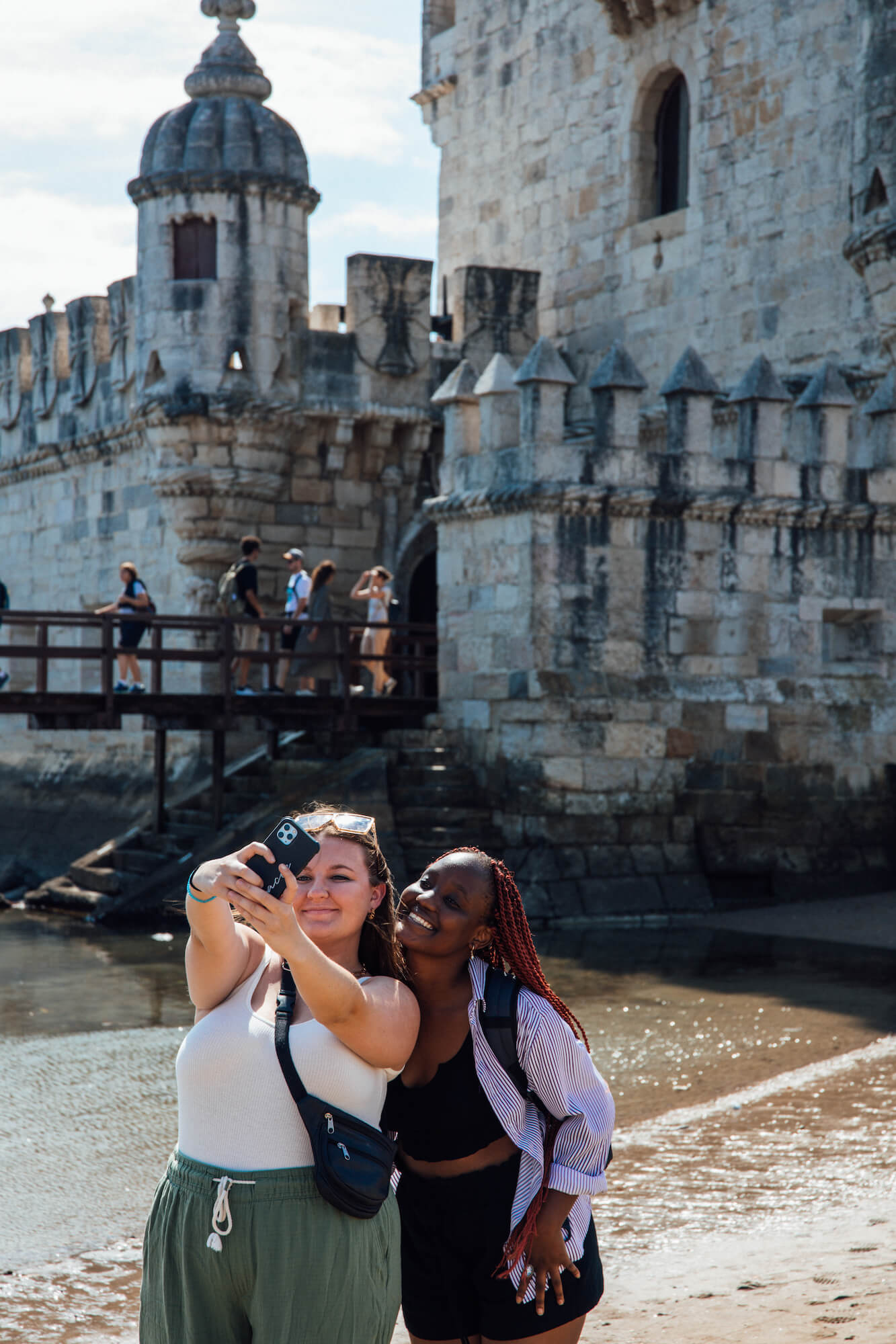 Two young women pose for a selfie on a beach in front of a stone, castle-like building. In the background, people enter and exit the building via a wooden bridge over water.