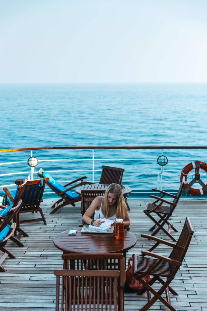 A young woman with long blonde hair sits at a wooden table on the deck of a ship, writing in a notebook. The blue ocean stretches to the horizon beyond the railing behind her. 
