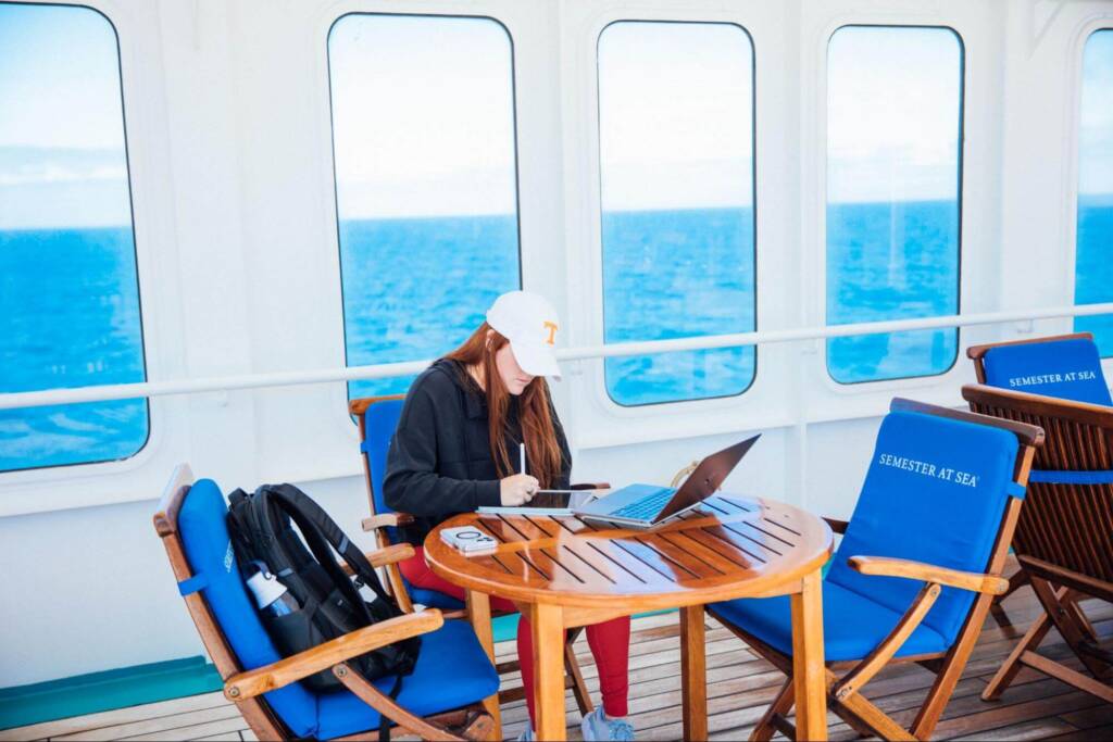 A student with long red hair and a baseball cap sits at a wooden table, working on an iPad with a laptop open in front of them. A very blue ocean is visible through porthole windows behind them.