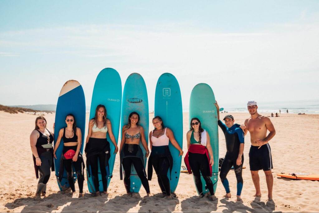 Eight people in bathing suits and wetsuits stand smiling on a sunny beach; five of them hold surfboards in different shades of blue.