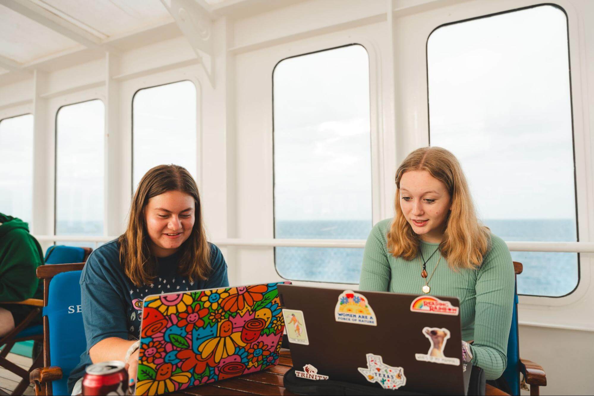 Two students sitting on a ship, working on laptops with colorful stickers, smiling and engaged in their work.