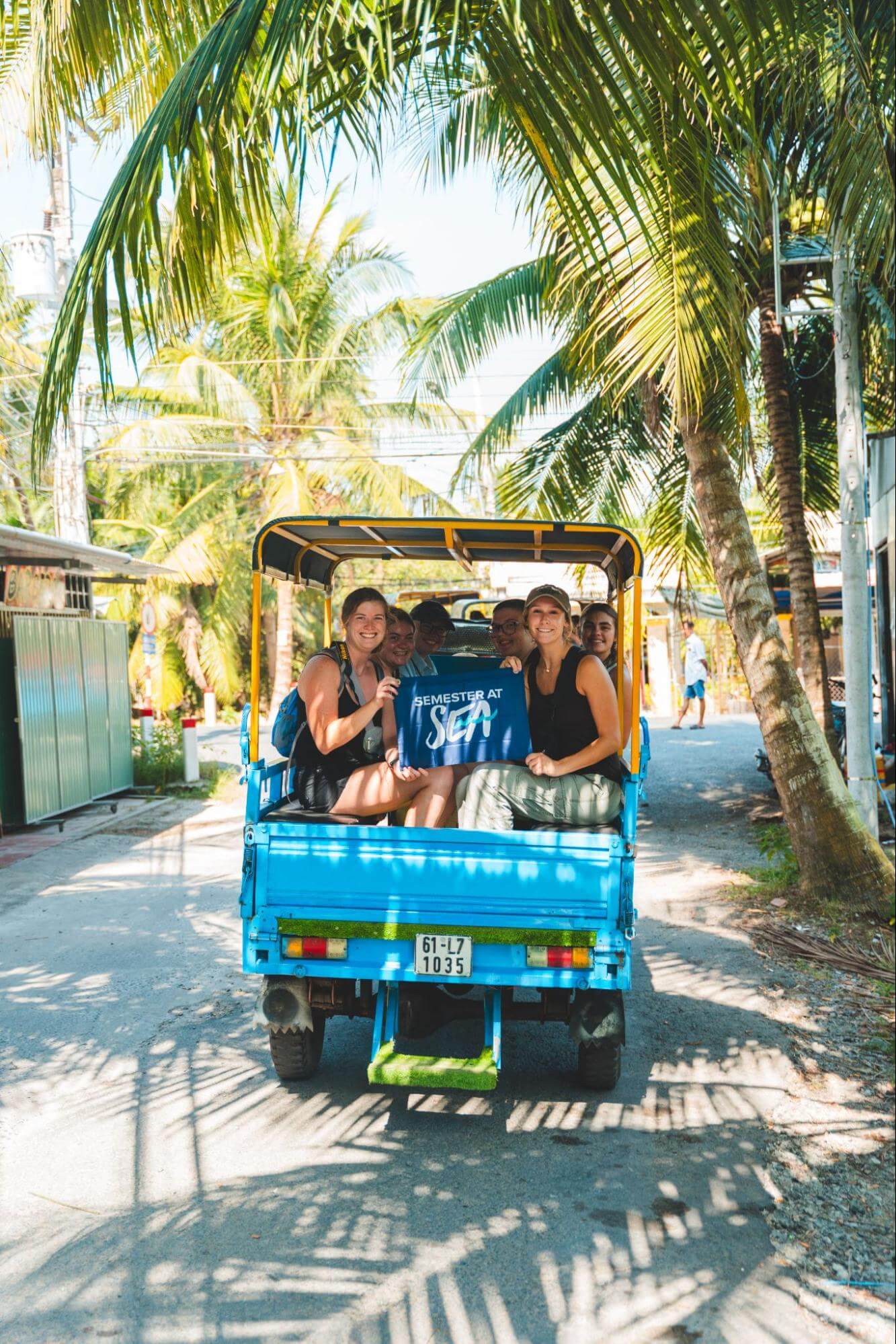 A group of students sit in the back of a bright blue truck with a covered bed, surrounded by palm trees, smiling and holding a blue flag that says “Semester at Sea.”