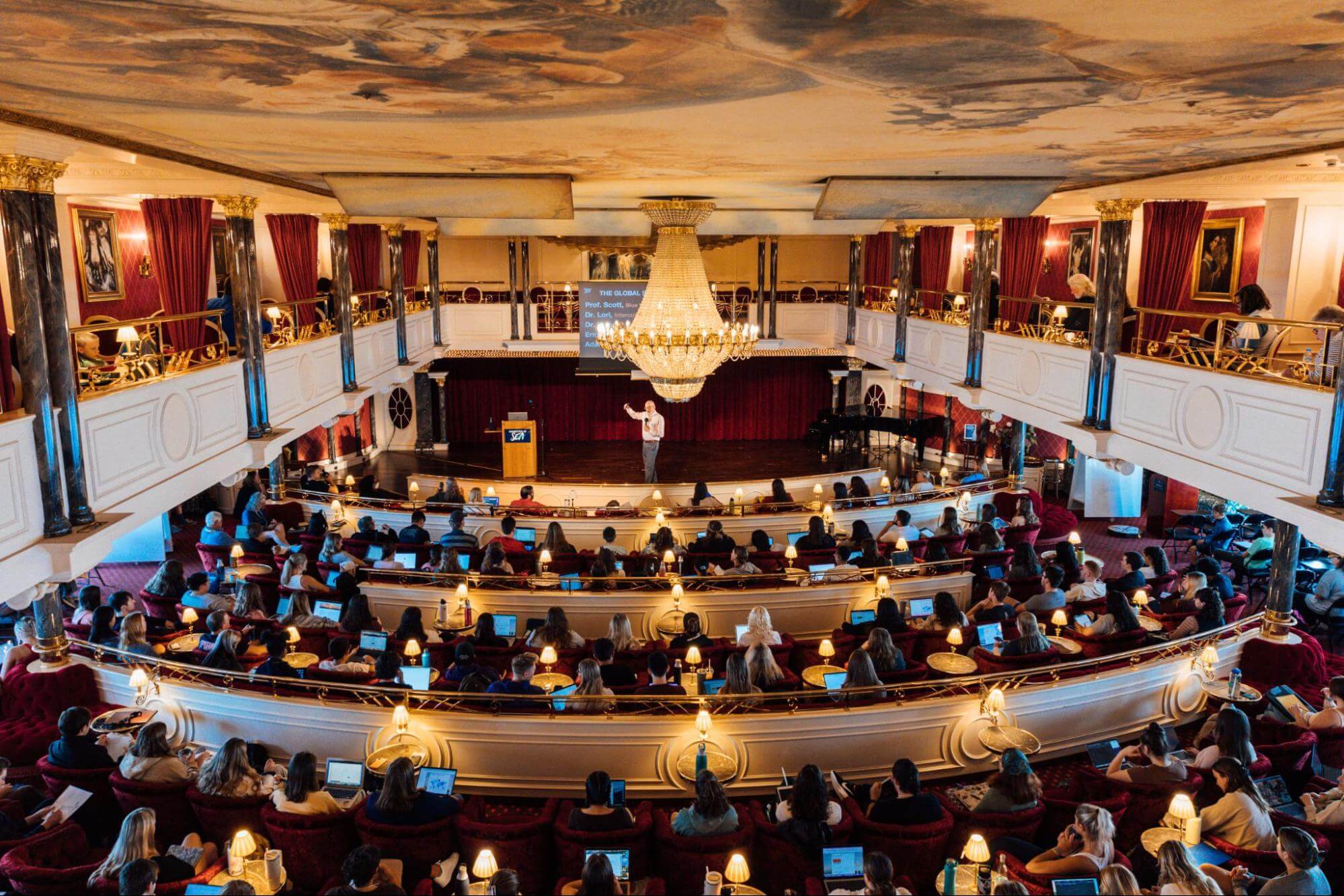 Students with laptops are seated in a large, opulent theater — complete with balconies, ceiling frescoes, and a chandelier — facing an instructor giving a lecture onstage.