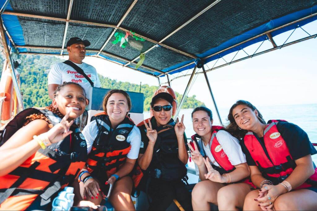 Five students wearing life jackets are seated in a covered boat, smiling and flashing peace signs to the camera. The boat operator stands at the helm behind them.