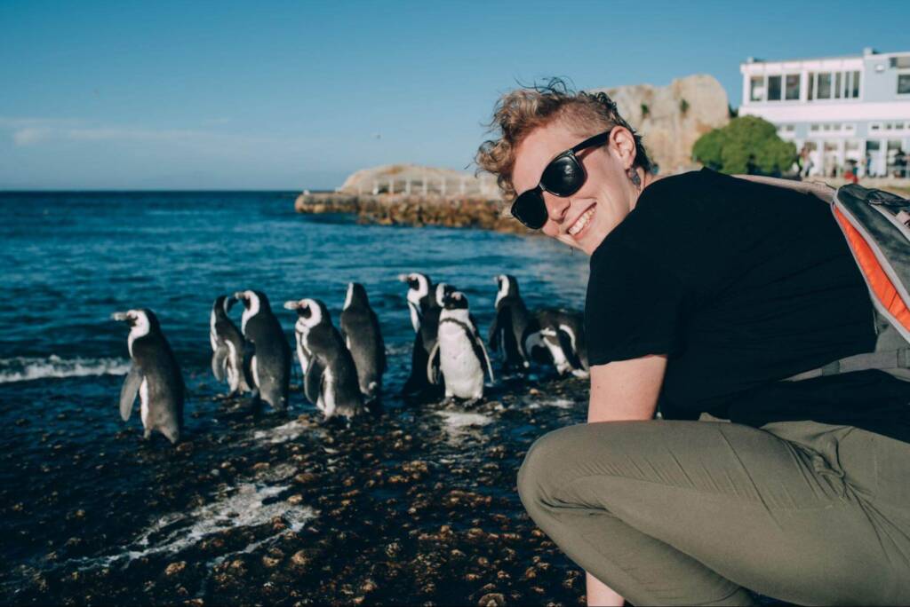 A young woman in sunglasses couches next to a flock of penguins on a rocky shoreline. She smiles at the camera as the penguins stand in the shallows, looking out toward open water.