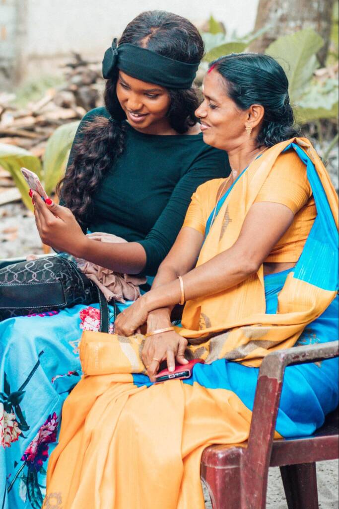 A young woman and an older woman are seated side-by-side; the young woman, in a green headband, shows the older woman something on her phone. The older woman, wearing a blue and orange sari, smiles at the phone screen.