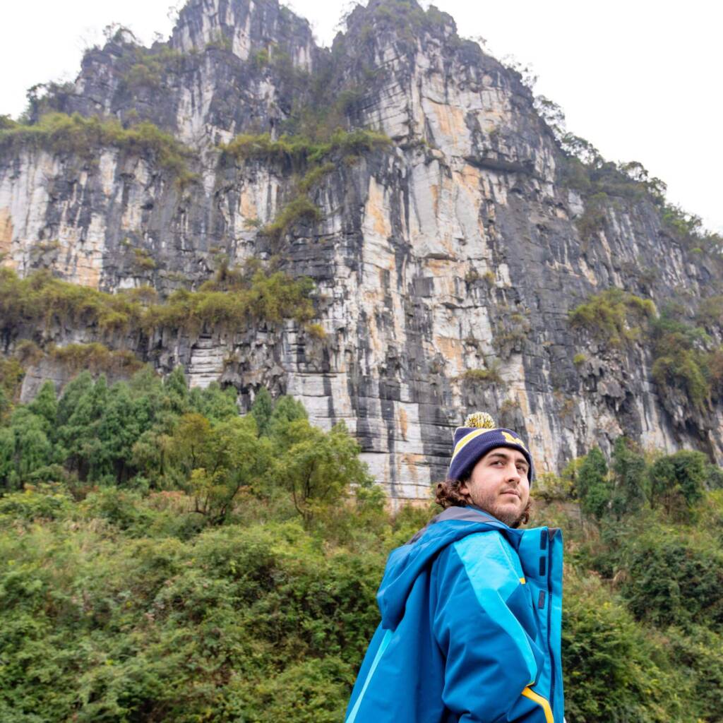 A young man in a blue jacket and navy blue beanie looks over his shoulder toward the camera, away from the sheer rocky cliff face that rises in front of him.