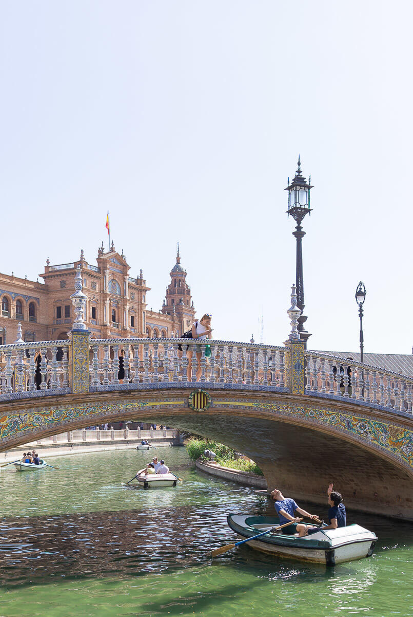 Two young men in a small rowboat in a city canal look up at a young woman as she takes their picture from a mosaiced stone bridge above them. An ornate building flying the Spanish flag rises behind her. 