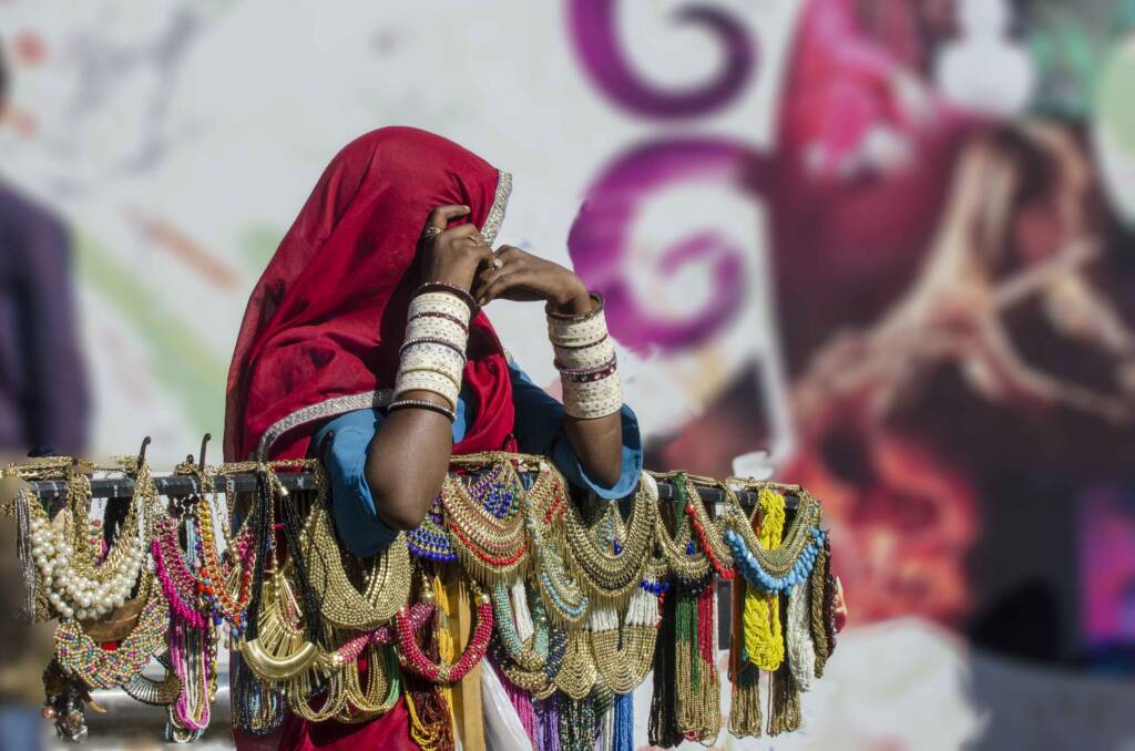 A woman wearing a red headscarf and stacks of bangles stands behind a street display of ornate beaded necklaces.