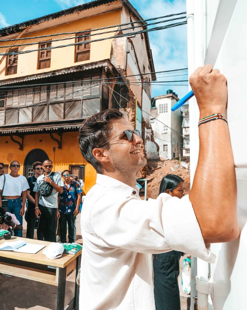 A man in a white shirt and sunglasses writes on the wall of a building with a blue marker, while a group of people look on behind him. 