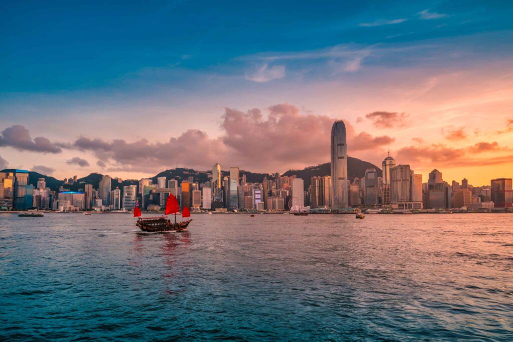A junk-style boat with bright red sails floats on a harbor against a backdrop of the Hong Kong skyline at sunset.