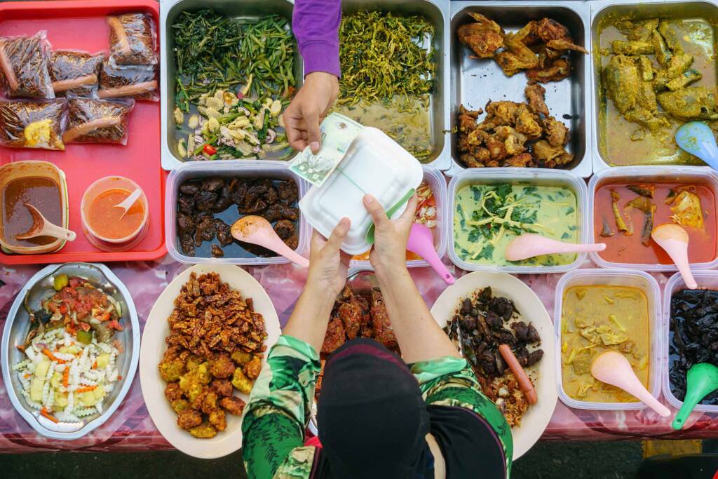 An overhead view of a person paying cash for a styrofoam takeout container over a table full of colorful dishes of food. 
