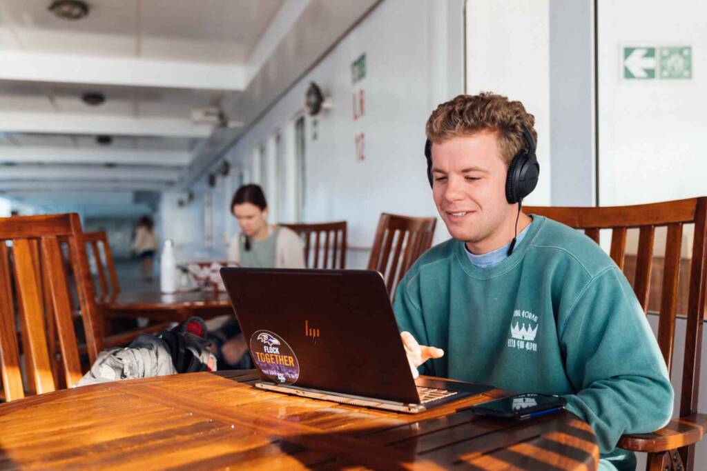 A young man in a teal sweatshirt sits at a wooden table in front of a laptop, wearing headphones and speaking on a virtual call.